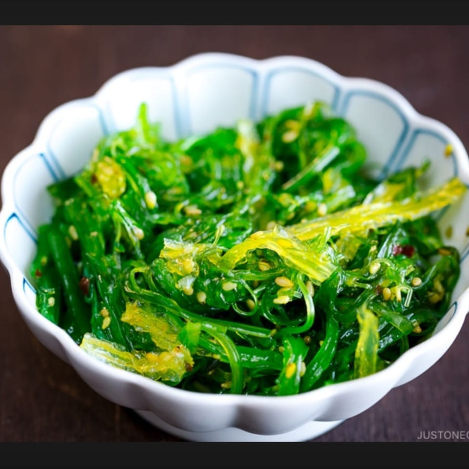 A white scalloped bowl filled with vibrant green seaweed salad, garnished with sesame seeds.