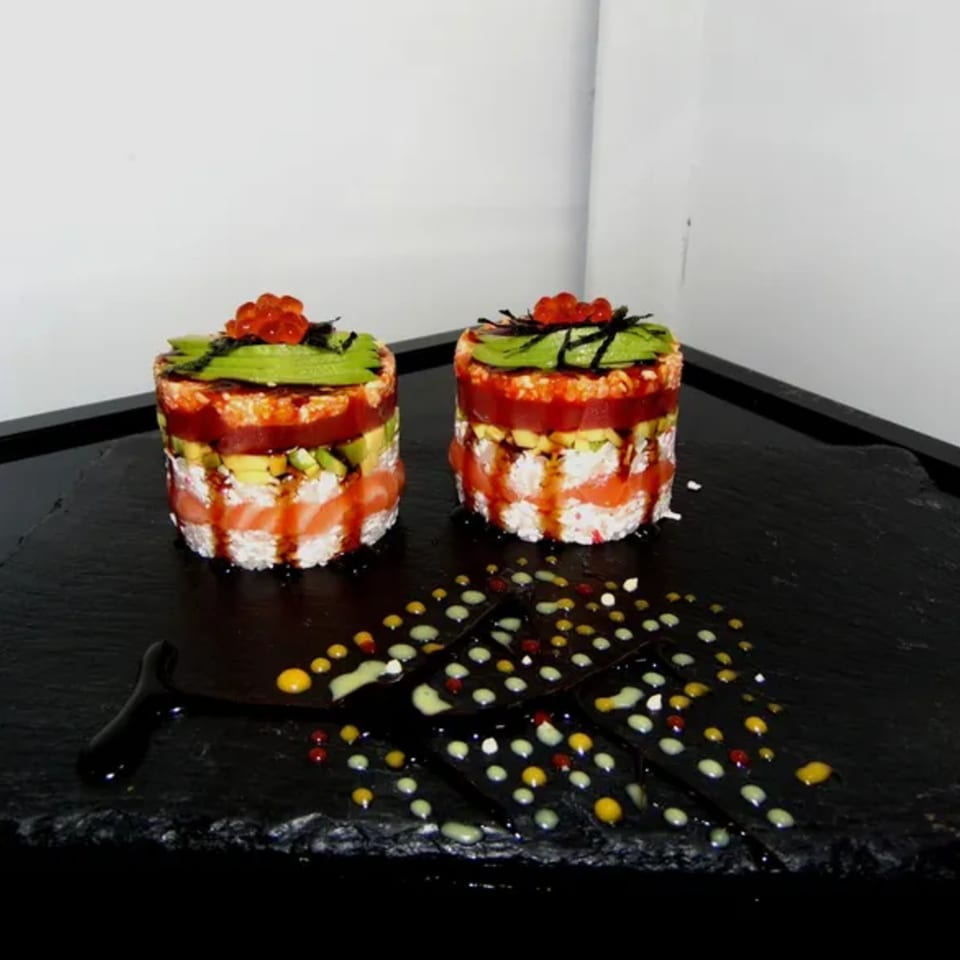 Two layered sushi towers with avocado, salmon, tuna, and roe, on a black slate.